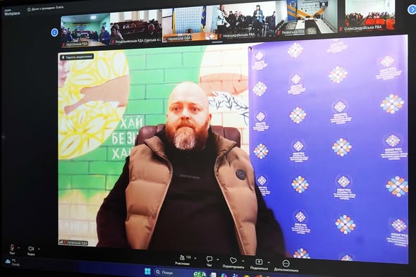 У центрі кадру — чоловік середнього віку з густою русявою бородою з сивиною. Він одягнений у чорну кофту та бежевий дутий жилет. Чоловік дивиться прямо в камеру.