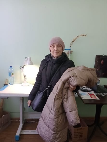 Одягнена в теплу чорну стьобану куртку та світло-рожеву (пудрову) в'язану шапку. Через плече у неї висить чорна сумка. Вона тримає в руках ще одну об'ємну довгу куртку бежевого або світло-коричневого кольору.