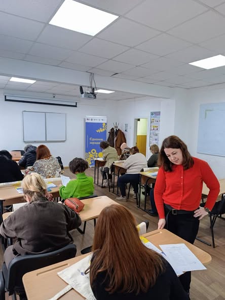У кімнаті перебуває група жінок різного віку. Більшість із них сидять за партами, розставленими рядами, і зосереджено працюють із паперами — ймовірно, заповнюють анкети або виконують навчальні завдання.