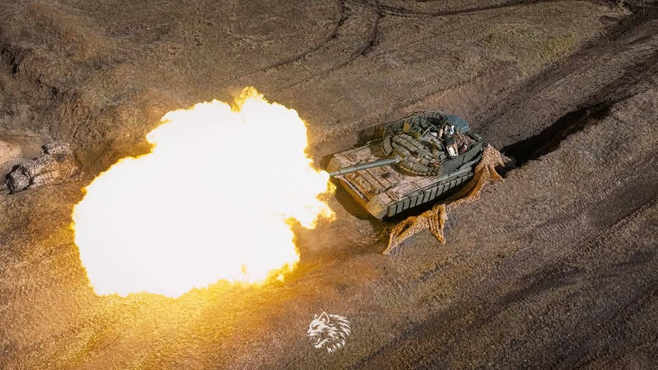 На знімку зафіксовано потужний момент пострілу танка з ракурсу зверху, де величезний вогняний спалах з дула контрастує з темною зораною землею та камуфляжною сіткою поруч