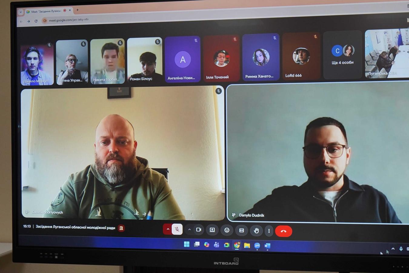 На моніторі марки «INTBOARD» відображено вікно онлайн-зустрічі в Google Meet, де учасники засідання Луганської обласної молодіжної ради, серед яких двоє чоловіків на передньому плані та декілька інших у мініатюрах зверху, обговорюють робочі питання в режимі відеоконференції.