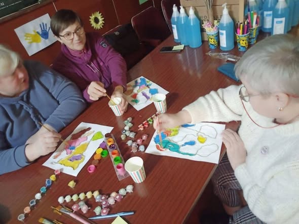 Три жінки різного віку зосереджено займаються арттерапією за великим столом, розфарбовуючи яскраві абстрактні малюнки фарбами за допомогою тонких пензлів та стаканчиків з водою