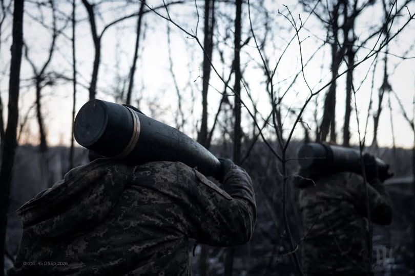 На фотографії в атмосфері сутінків або раннього світанку зображені двоє військових у камуфляжній формі, які йдуть крізь густий ліс із голими деревами. Кожен із них несе на плечі великий артилерійський снаряд або тубус від нього