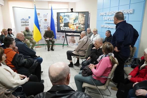 Фото зроблено з ракурсу ззаду глядачів, що сидять у колі. У центрі уваги — телевізійний екран із зображенням, що нагадує зону відчуження. Ліворуч стоять два прапори: державний прапор України та синій прапор із гербом. Спікер у військовій формі сидить із нотатником і звертається до аудиторії. Атмосфера офіційна та стримана.
