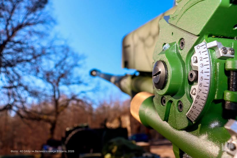 На зображенні представлено крупний план механізму прицілювання сучасної артилерійської установки зеленого кольору зі шкалою та цифрами, на тлі якої в розмитій перспективі видно ствол гармати, спрямований у безхмарне блакитне небо над лісистою місцевістю.