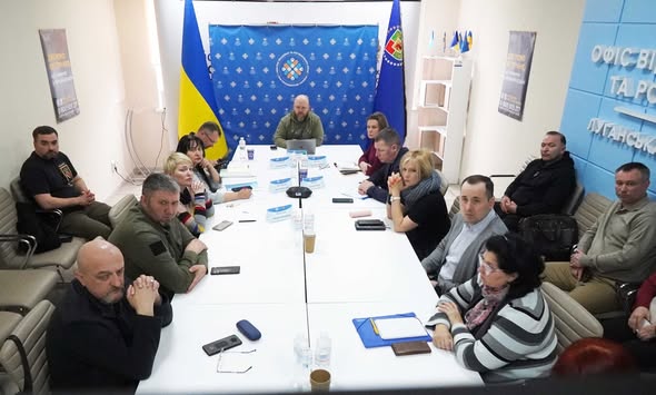 Група людей у цивільному та військовому одязі проводить офіційне засідання за великим білим столом у кабінеті, прикрашеному державною символікою України та інформаційними стендами.