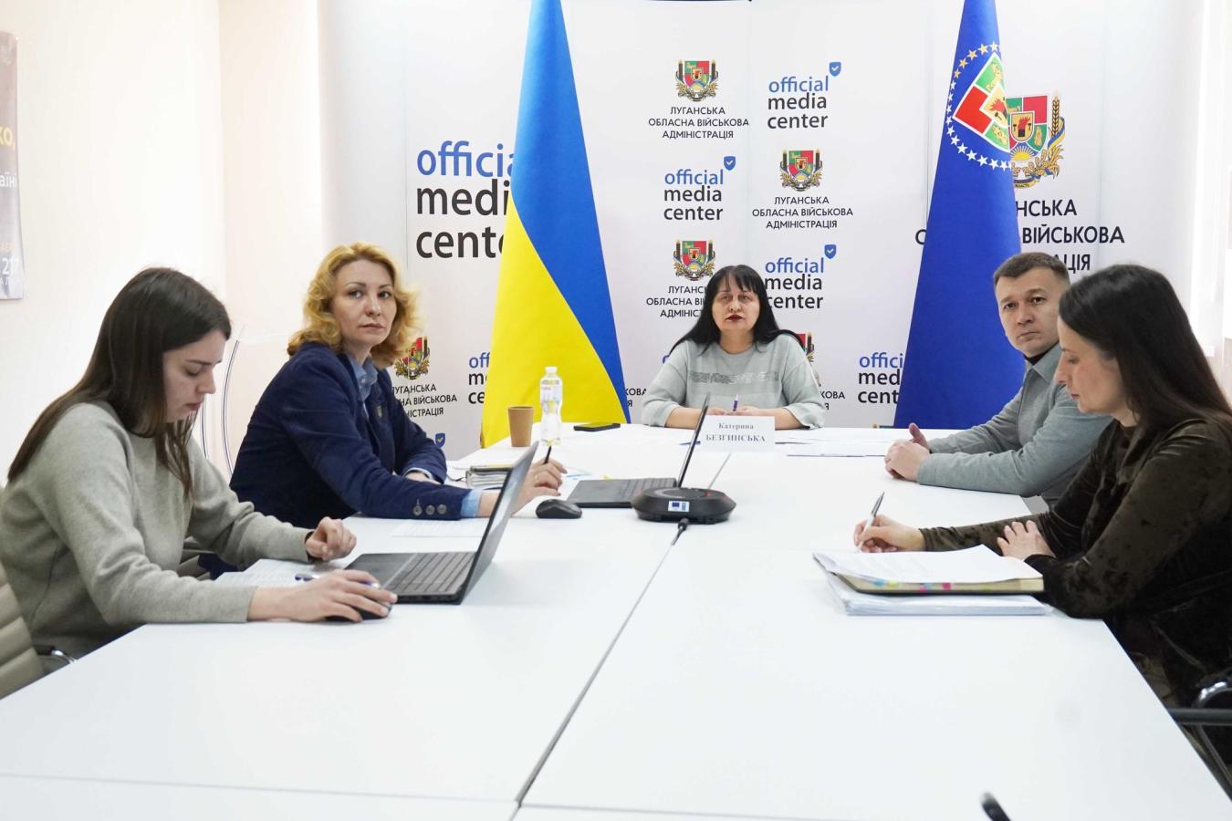 П’ятеро учасників засідання в «Official Media Center» Луганської обласної військової адміністрації сидять за білим Т-подібним столом із ноутбуками та паперами на фоні прапорів України та брендованих банерів.