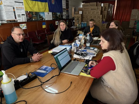 Група людей зосереджено працює за довгим дерев'яним столом, заставленим ноутбуками та документами, у приміщенні з українською символікою та численними коробками гуманітарної допомоги на задньому плані