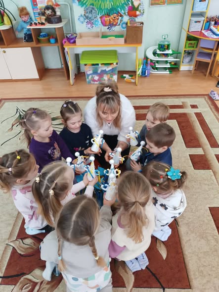  Вихователька та група з дев'яти маленьких дітей (хлопчиків та дівчаток) сиділи колом на килимі. Усі вони тримають у руках саморобні іграшки — білих паперових янголів з німбами.