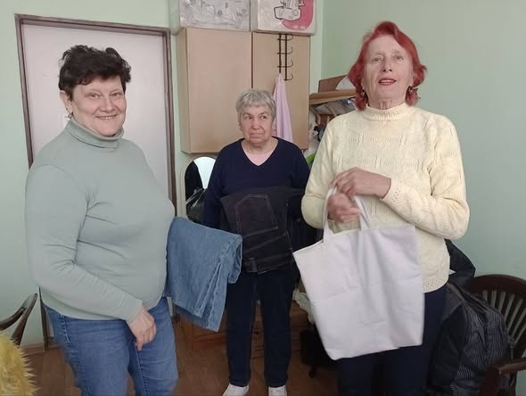 На знімку зображені три жінки старшого віку, які стоять у приміщенні. Вони дивляться в об'єктив і злегка посміхаються, створюючи атмосферу доброзичливості та взаємодопомоги