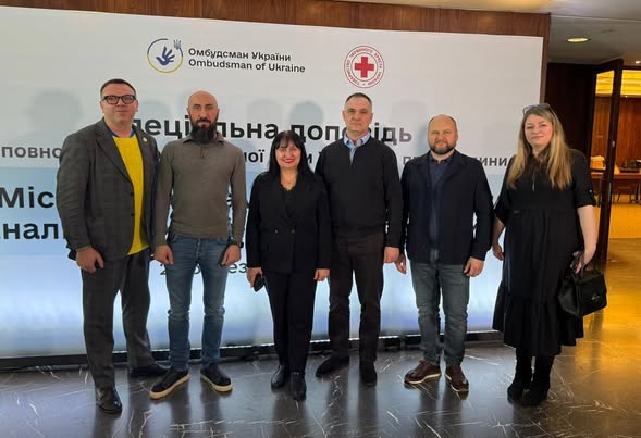 Група з шести офіційно вдягнених людей — чотирьох чоловіків і двох жінок — позують для спільного фото на фоні банера з логотипами Омбудсмена України та Червоного Хреста під час презентації спеціальної доповіді.
