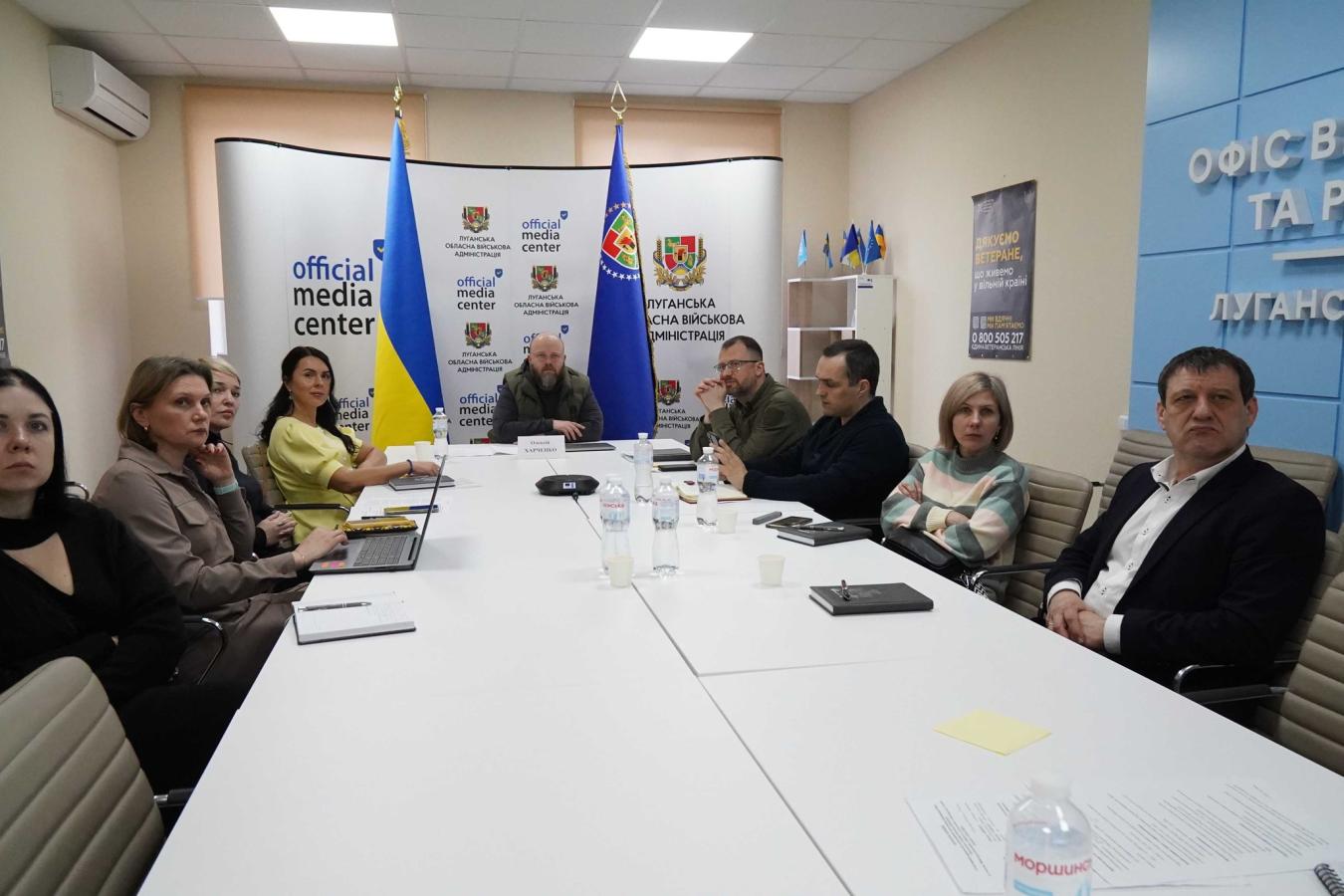 Група посадовців Луганської обласної державної адміністрації та представників медіацентру проводить робочу нараду за великим білим столом в офіційному кабінеті, прикрашеному державними прапорами та символікою адміністрації.