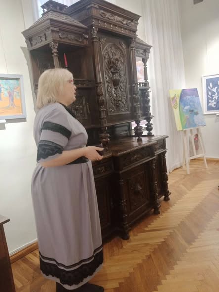 Жінка у довгій сірій сукні з мереживом проводить екскурсію у світлій музейній залі з паркетною підлогою, розповідаючи про масивний старовинний буфет із темного дерева, прикрашений складним художнім різьбленням.