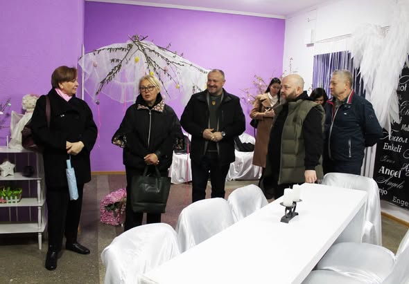 Група людей оглядає світлу кімнату з фіолетовими стінами, яка прикрашена до свята або тематичного заходу. У центрі — довгий білий стіл зі стільцями в білих чохлах. На задньому плані видно декоративне дерево з писанками та ангельські крила.