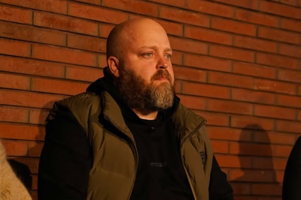 Портрет чоловіка з густою бородою, який сидить на фоні цегляної стіни. Чоловік одягнений у чорне худі та коричневий жилет, його погляд спрямований убік. Тепле вечірнє освітлення створює глибокі тіні.