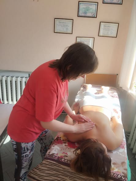 На знімку зображена масажистка (жінка у червоній футболці та спортивних легінсах), яка проводить процедуру. Вона зосереджено працює над областю спини та лопаток пацієнтки.