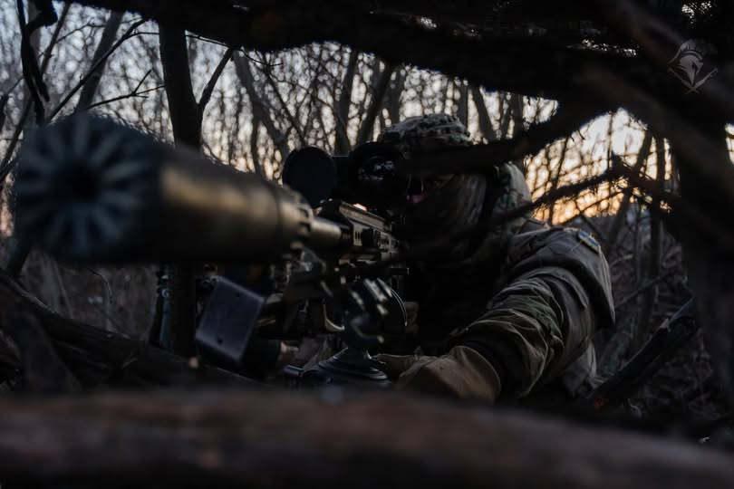 Український військовий у повному спорядженні та балаклаві веде спостереження через оптичний приціл гвинтівки з глушником, перебуваючи на замаскованій позиції серед густих гілок та лісової рослинності.