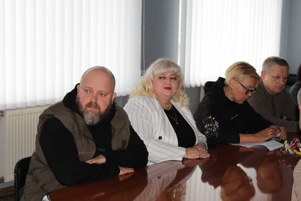 Четверо людей сидять за довгим полірованим столом під час зустрічі або наради. На задньому плані — вікна з білими вертикальними жалюзі. Учасники виглядають зосередженими, панує робоча атмосфера.