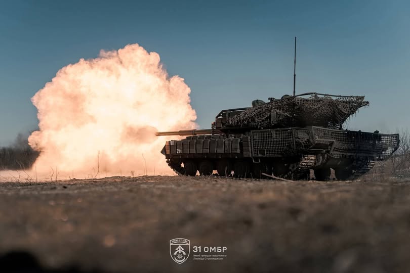 На фото зображено український танк (імовірно Leopard 1A5 або модернізований Т-64), оснащений додатковим захистом. Зокрема, помітно маскувальну сітку на башті та навісні елементи динамічного захисту на корпусі й бортах.