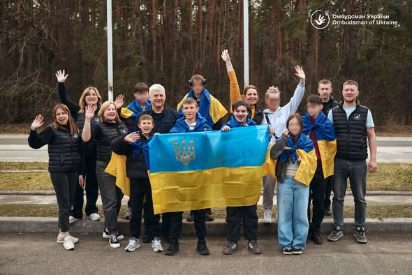На знімку зображено близько 15 осіб — дітей різного віку та дорослих, які супроводжують їх. Вони стоять щільною групою на узбіччі дороги на фоні густого соснового лісу. Атмосфера сповнена радості, полегшення та патріотичного піднесення.