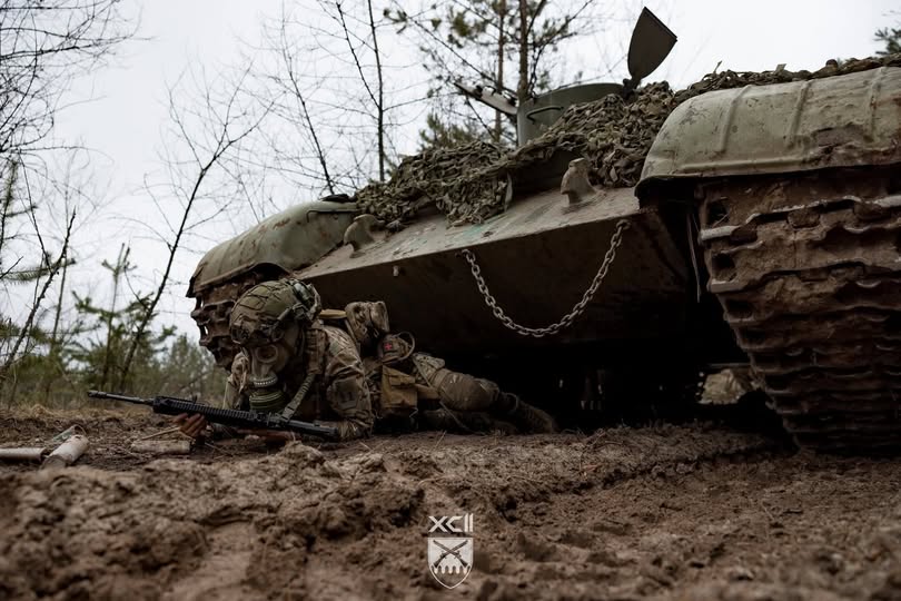 Український військовослужбовець у повному бойовому спорядженні та протигазі веде спостереження з автоматичною зброєю, перебуваючи в укритті безпосередньо під корпусом замаскованого бронеавтомобіля на ґрунтовій дорозі в лісистій місцевості.