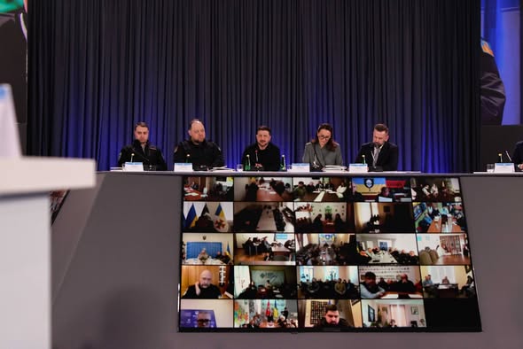 На фоні темних штор за довгим столом під час офіційного заходу сидять п'ятеро представників влади, серед яких у центрі Володимир Зеленський, а перед ними на великому похилому екрані відображається мозаїка з десятків вікон відеозв'язку з іншими учасниками конференції.