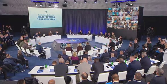 У великому сучасному залі під час заходу «Діалог громад з Парламентом» численні учасники в діловому вбранні розмістилися за напівкруглими столами навколо центральної восьмикутної трибуни, а на великих екранах відображаються назва панельної дискусії та десятки вікон із відеозв’язком дистанційних спікерів.