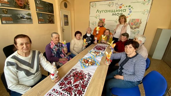 Група жінок старшого віку зібралася за довгим столом, застеленим вишитим рушником і прикрашеним крашанками, тримаючи в руках саморобні великодні сувеніри на тлі плаката з написом «Луганщина моя»