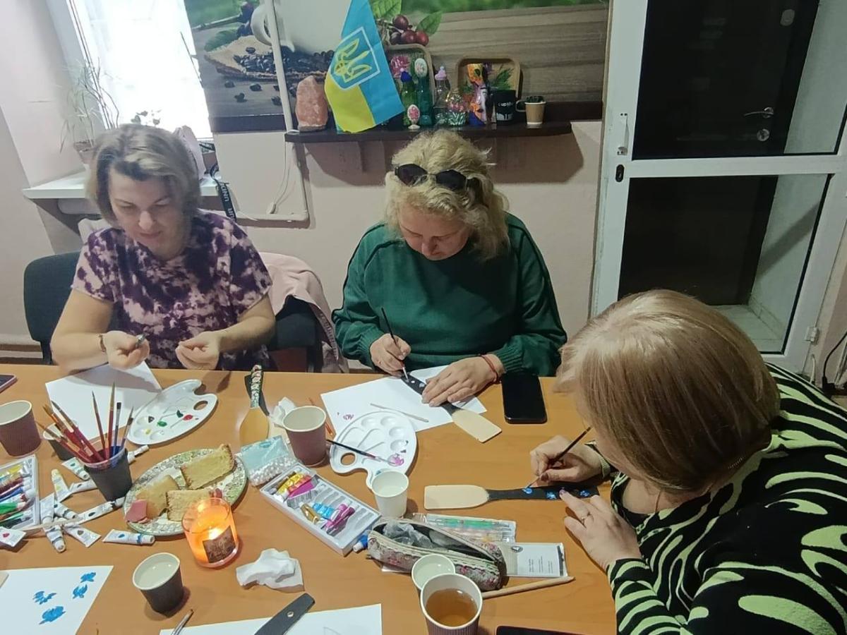 Три жінки зосереджено працюють за столом під час творчого майстер-класу в гуманітарному осередку, розписуючи дерев’яне кухонне начиння традиційними орнаментами в оточенні художнього приладдя, святкової випічки та державної символіки.