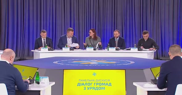 На тлі масивних синіх штор п'ятеро спікерів у діловому вбранні сидять за довгим білим столом із мікрофонами, проводячи панельну дискусію «Діалог громад з Урядом», назва якої яскраво світиться на жовтому екрані перед столом.