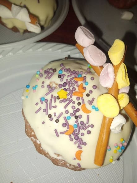 Святковий кекс, покритий білою глазур'ю та яскравою декоративною посипкою, прикрашений оригінальною їстівною композицією у формі вербових котиків, виготовленою з хрусткої соломки та кольорового маршмелоу.