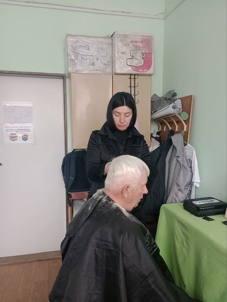 На світлині жінка у темному одязі стриже літнього чоловіка в накидці у скромно облаштованому приміщенні з шафами та вішалкою для одягу на фоні.