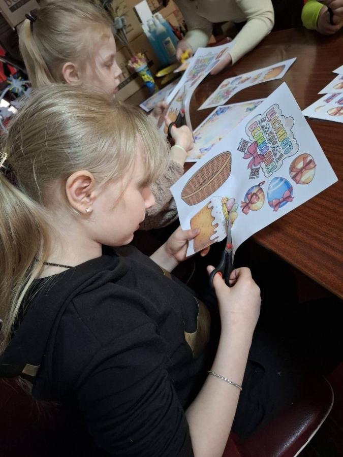 Дві дівчинки зосереджено вирізають ножицями яскраві великодні ілюстрації із зображеннями пасок, кошиків та крашанок з аркушів паперу, готуючи саморобні прикраси до свята під час творчого заняття