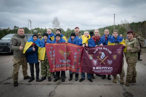Група українських захисників та цивільних позує для спільного фото просто неба, тримаючи великі бордові прапори з символікою 8-го армійського корпусу та Десантно-штурмових військ, при цьому деякі учасники загорнуті в національні синьо-жовті стяги.