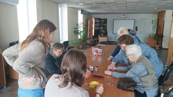 На фото зображено групу жінок різного віку, які зібралися навколо довгого дерев’яного столу в просторому приміщенні. Вони зосереджено займаються спільною справою, схожою на майстер-клас із рукоділля або волонтерську діяльність.