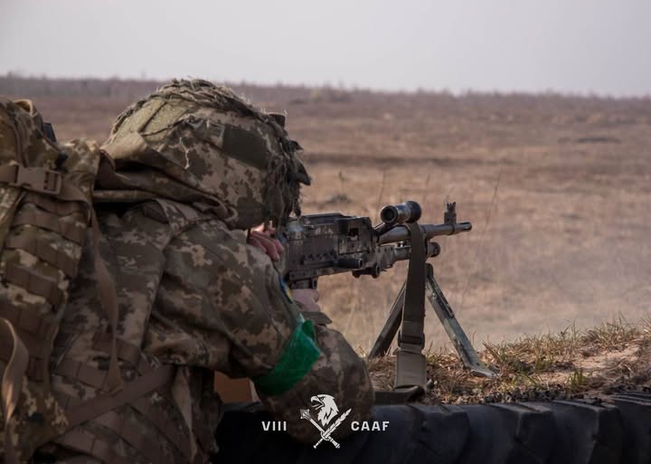 Військовослужбовець, знятий зі спини та збоку (у профіль). Він схилився над зброєю, зосереджено дивлячись у приціл.