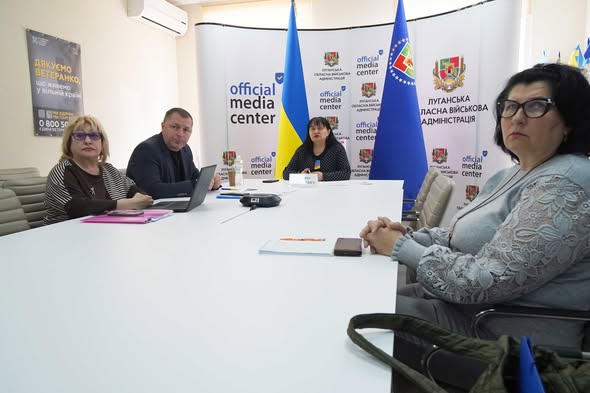Четверо людей сидять за білим столом у приміщенні «Official Media Center» Луганської обласної військової адміністрації під час проведення брифінгу або робочої зустрічі на фоні державних прапорів.