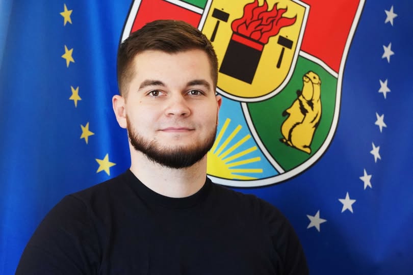 Портрет молодого чоловіка з темною бородою у чорній футболці на фоні синього прапора з жовтими зірками та великого кольорового герба Луганської області із зображенням бабака та сонця.
