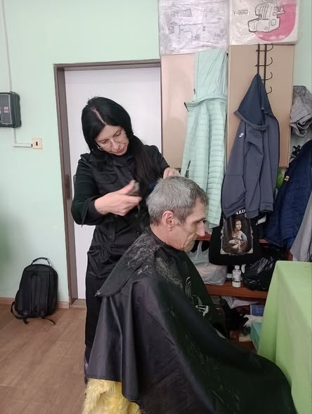 Жінка-перукар у чорному одязі стриже сивого чоловіка, накритого темним пеньюаром, у простому світлому приміщенні, де на фоні видно двері, вішалку з одягом та полиці з коробками
