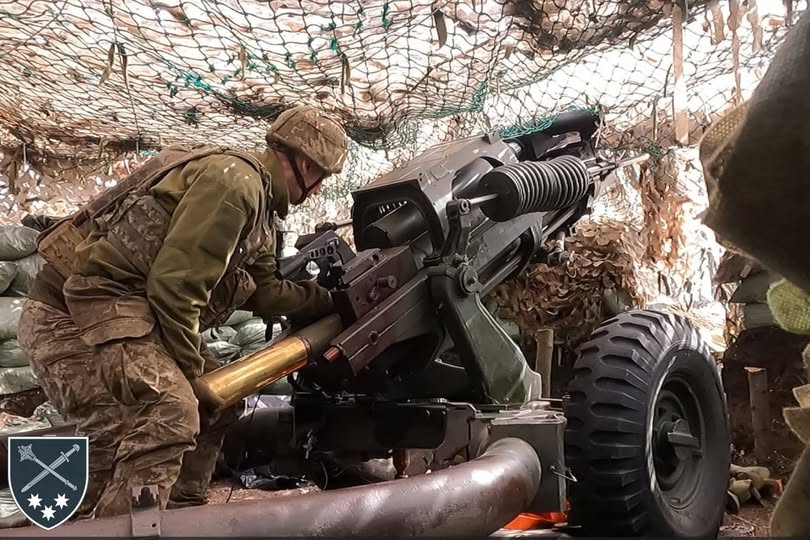 На фотографії український військовослужбовець у камуфляжній формі та шоломі, перебуваючи в укріпленій позиції під маскувальною сіткою, заряджає артилерійську гармату великим снарядом