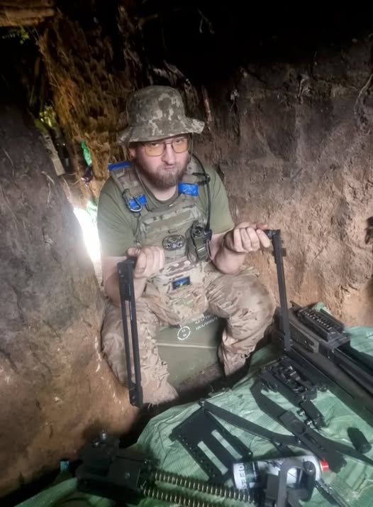 На фото зображений бородатий український військовослужбовець в окулярах, камуфляжній панамі та плитоносці, який сидить у вузькому земляному окопі та тримає в руках деталі від розібраного великокаліберного кулемета, розкладеного перед ним на підстилці.
