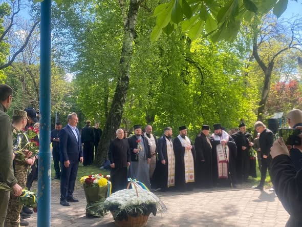 На фотографії шеренга православних священнослужителів у темному церковному вбранні та чоловік у синьому костюмі стоять біля великих кошиків із квітами під час офіційного пам'ятного заходу в оточенні густої зелені парку.