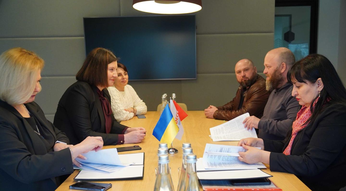 За довгим дерев'яним столом у сучасному офісному приміщенні з вимкненим екраном на стіні проходить офіційна зустріч шести осіб, де три жінки ліворуч та двоє чоловіків із жінкою праворуч обговорюють документи, сидячи навпроти маленьких настільних прапорців України та організації «Десяте квітня»