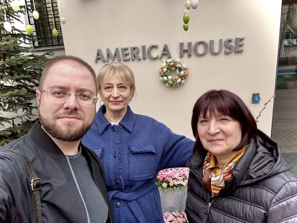 На фоні входу до будівлі з написом «AMERICA HOUSE», прикрашеного великоднім вінком та підвісними декоративними яйцями, троє усміхнених людей — чоловік в окулярах та чорній куртці і дві жінки в синьому пальті та темному пуховику — роблять спільне селфі.