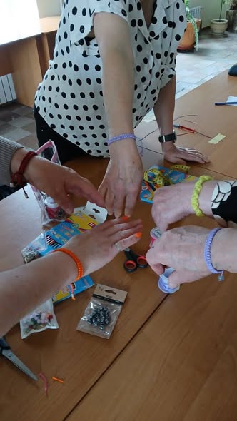 На дерев'яному столі, заваленому пакетиками з бісером, котушками ниток і ножицями, кілька людей у різнокольорових плетених браслетах простягають руки до центру, демонструючи результати спільного майстер-класу з рукоділля.