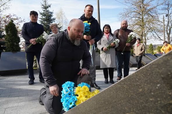 На передньому плані чоловік схилився на одне коліно біля кам'яної плити меморіалу, покладаючи квіти синьо-жовтих кольорів, тоді як позаду стоїть група людей із білими та синьо-жовтими букетами, вшановуючи пам'ять загиблих