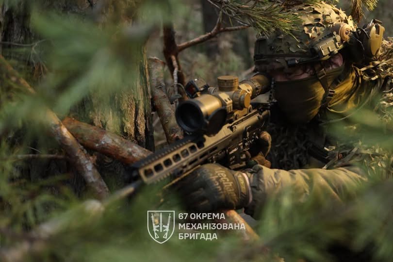 Український військовий 67-ї окремої механізованої бригади у повному бойовому спорядженні та камуфляжі прицілюється з гвинтівки з масивним оптичним прицілом, знаходячись у густих лісових хащах.