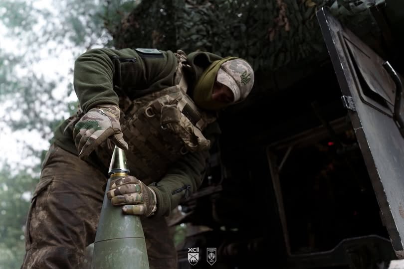 Український військовий у тактичному спорядженні та балаклаві зосереджено готує артилерійський снаряд до пострілу на фоні замаскованої бойової машини в лісистій місцевості