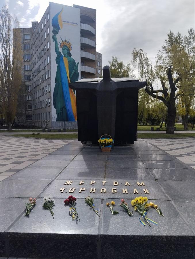 На передньому плані фотографії розташовано гранітний пам'ятник «Жертвам Чорнобиля» із покладеними біля підніжжя квітами, а на задньому плані височіє багатоповерхівка з великим муралом, де Статуя Свободи одягнена у синьо-жовтий прапор та вінок із соняшників.