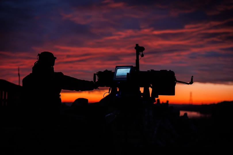 На фотографії зображено темний силует військового оператора, який працює з автоматизованою установкою з увімкненим монітором на тлі мальовничого багряно-червоного неба під час заходу сонця